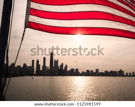 A beautiful picture of an American Flag rippling in the wind over the Chicago skyline at sunset with building silhouettes and sun reflecting on the water of Lake Michigan.