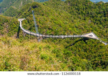 High-angle shot of Langkawi Sky Bridge, a curved bridge with a triangular viewing platform at each end of the walkway, suspended by cables from one pylon at the peak of Gunung Machinchang, Malaysia.
