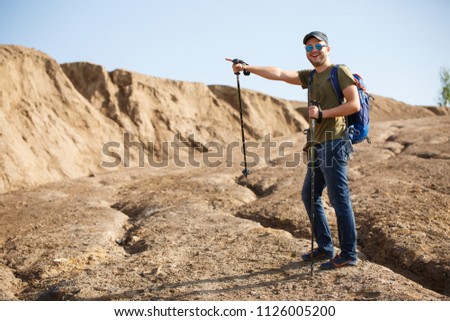 Picture of athlete man with backpack and walking sticks pointing with hand to side