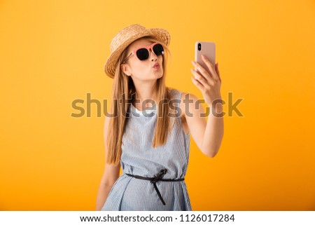 Lovely young blonde woman in summer hat and sunglasses taking a selfie isolated over yellow background