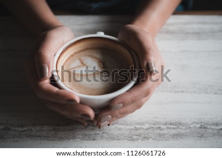 Top view image of a woman's hands holding a cup of hot latte coffee on table in cafe