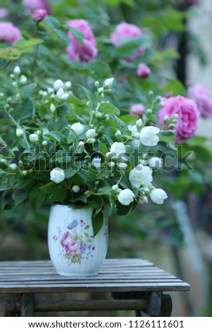 Floral scene in garden with bouquet of jasmine in vintage porcelain jar on old aged wooden stool on bush of roses background, vertical photo, outdoors and space, natural evening light, vertical photo 