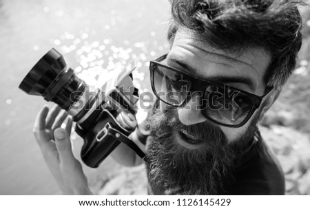 Man with beard and mustache wears sunglasses, water surface on background. Hipster on smiling face holds old fashioned camera. Tourist photographer concept. Guy shooting nature near river or pond.