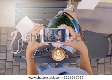 Modern woman make photo with coffee, bouquet with pink pionies, spectacles and little pink backpack in outdoor restaurant. top view