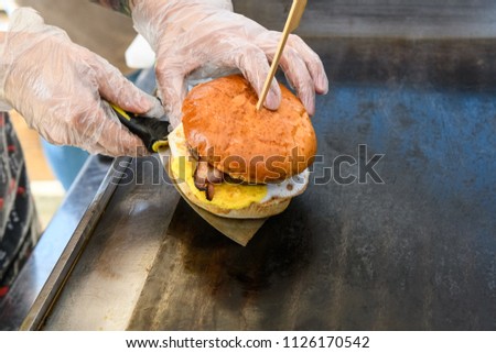 The chef transfers a delicious burger from a metal spatula with a white bun, scrambled eggs and bacon, and a grill for cooking the dish.
