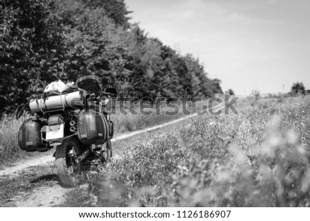 Motorcycle alone traveler with suitcases standing on field dirt road, forest and flowers, off road trip concept adventure, black and white
