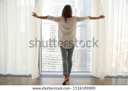 Rear view at rich woman standing looking out of full-length window of luxury modern apartment or hotel room opening curtains in the morning enjoying sunlight and city skyscrapers view feeling happy Royalty-Free Stock Photo #1126189880