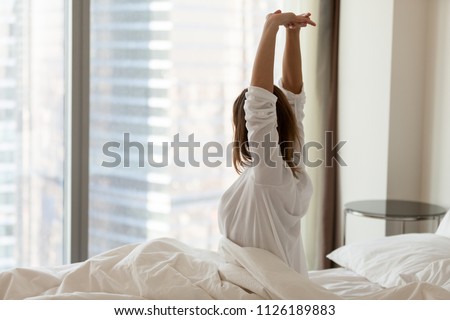 Woman waking up happy stretching sitting on comfortable bed looking out of big skyscraper window in modern hotel bedroom enjoying good morning and city view starting new day, wellbeing concept Royalty-Free Stock Photo #1126189883