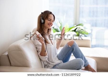 Smiling young woman in headphones listening to good music playing via smartphone application relaxing at home, happy millennial girl enjoying favorite songs podcast holding phone wearing earphones