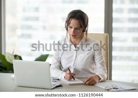 Serious businesswoman in headphones watching webinar on laptop making notes, learning studying computer course, making call, participating online conference, interpreter translating training class Royalty-Free Stock Photo #1126190444