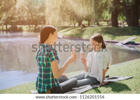 A picture of mom and daughgter sitting on blanket near lake. They are holding ice creams in hands. Girl is looking at ice cream. Woman is looking at child.