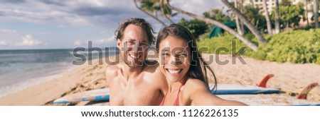 Beach vacation selfie couple. Happy young interracial friends taking photo with phone after surfing class on tropical Hawaii beach. Banner panorama. Travel vacation destination watersports.