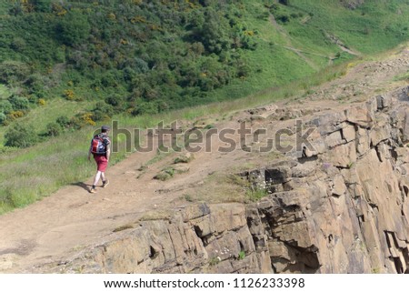 Man in shorts and backpack walking along a rocky plateau or ridge with wild gorse in background     