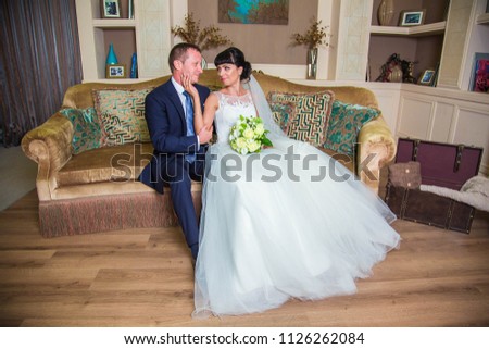 Bride in white dress, stroking groom's chin and looking at him, in luxurious room