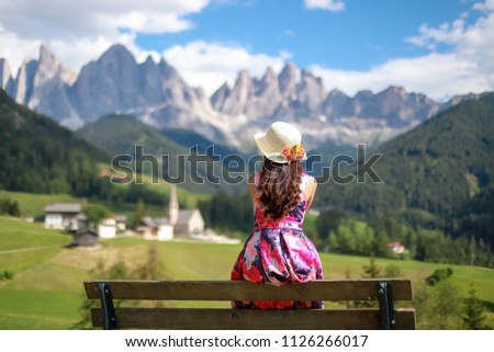 Woman sitting on bench relaxing her holiday travel to Dolomites in Italy. she wearing pink dress and straw hat with flower Looking to beautiful mountain peaks and the church of Santa Maddalena 