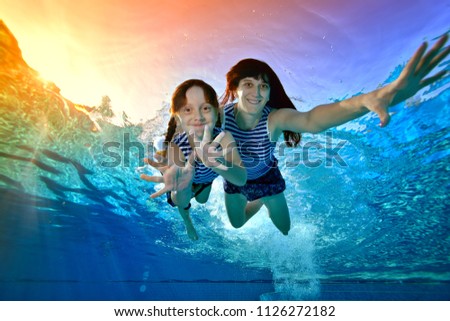 Happy mother with a little daughter swim underwater in striped bathing suits against the tropical sunset, look at the camera and smile. Portrait. Bottom view. Underwater photography.