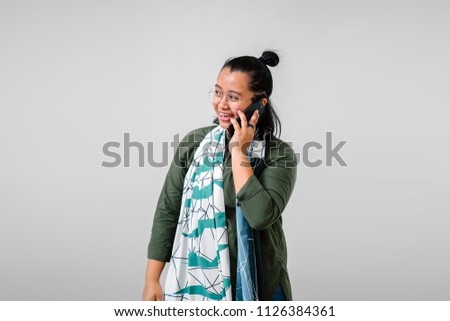 A portrait of a Malay woman smiling as she talks animatedly on her smartphone against a white background (studio). She is dressed like a creative sort in an olive green shirt and a colorful scarf. 