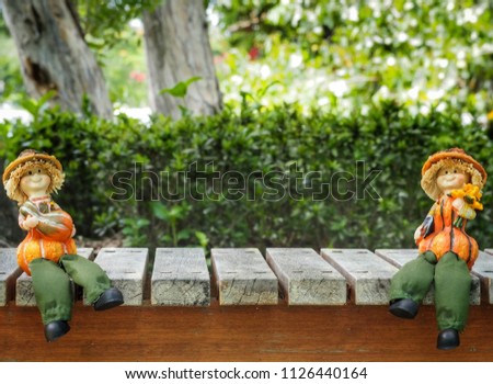 Two smmall farmer dolls sitting on wooden benches on the left and right sides of the picture. It has a green bushy background and has a central area for text input.