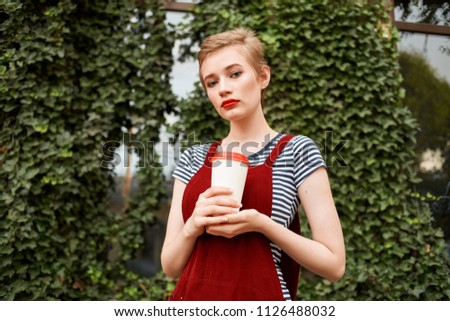 woman holding a light glass in her hand with a red lid and bushes in the background                            