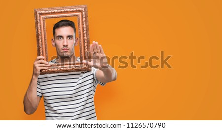 Handsome young man looking through vintage art frame with open hand doing stop sign with serious and confident expression, defense gesture