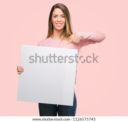 Beautiful young woman holding advertising banner with surprise face pointing finger to himself