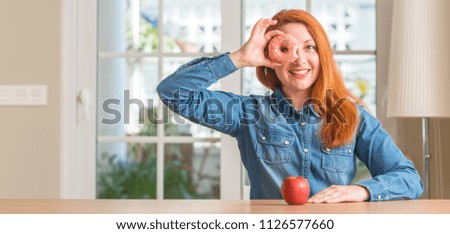 Redhead woman chooses between apple and donut with happy face smiling doing ok sign with hand on eye looking through fingers