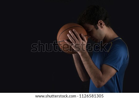 professional basketball player wearing sportswear and holding the ball leaning his head on it and making a wish, standing on the dark on a black background