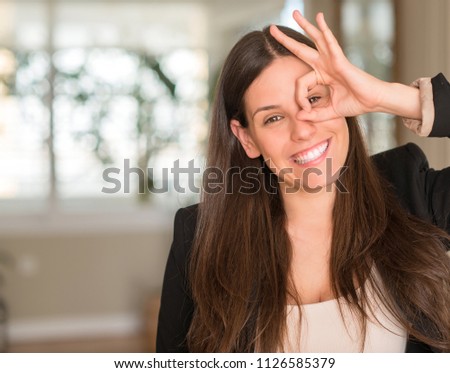 Young beautiful woman at home with happy face smiling doing ok sign with hand on eye looking through fingers