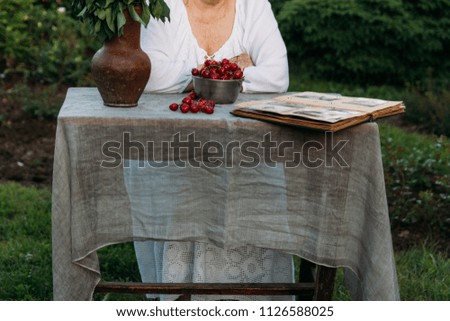 grandmother sitting in a chair at the table on which the cherry is lying, in the garden and watching an old album with photos, recounts and remembers the family's story. grandmother in a white vintage