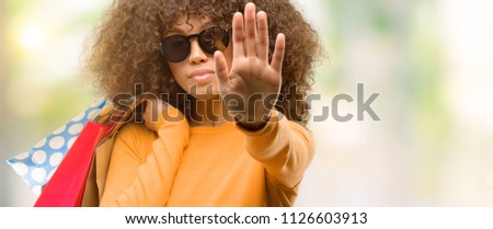 African american woman holding shopping bags with open hand doing stop sign with serious and confident expression, defense gesture