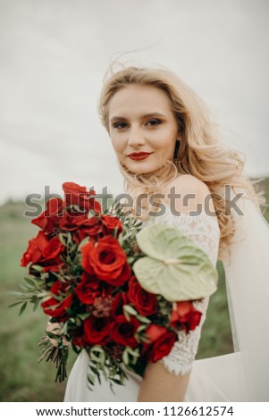 The joyful stylish beautiful fabulous happy blonde bride with the stylish bouquet is tenderly hugging flowers on the road in the countryside during the cloudy sunset.