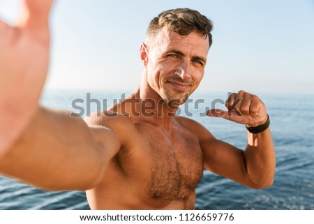 Smiling handsome shirtless sportsman taking a selfie and pointing finger at himself while standing at the beach