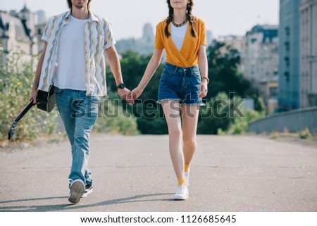 Young and happy man with guitar walking and holding hand of girl musicians in city
