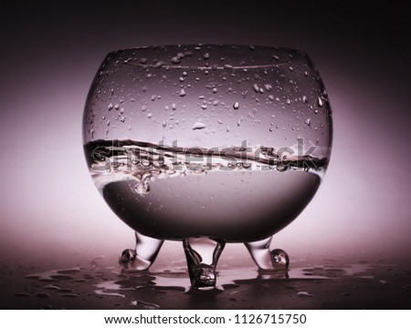 Fishbowl aquarium with water wave and bubbles in it on purple background with backlight. Water spilled onto the table.