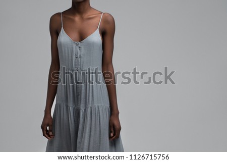 Black girl standing in blue-grey sundress on grey background. Her body is skinny and fit.