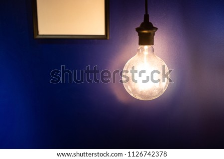 Big glowing light bulb against blue wall and picture frame with copy space