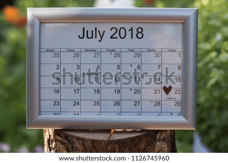 Close-up: calendar of the month July 2018 is in a silver frame on a wooden log among a summer green garden. Concept: Parents' day card.