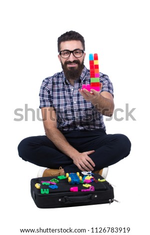 Unstable man build a tower of cubes raising it showing with a smile, sitting on the floor crossed legs, isolated on a white background.