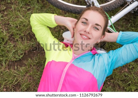 Young woman in sports wear relaxing in park after riding a bicycle. Fit, slim brunette woman with cheerful smile. Summer weather, outdoors leisure.