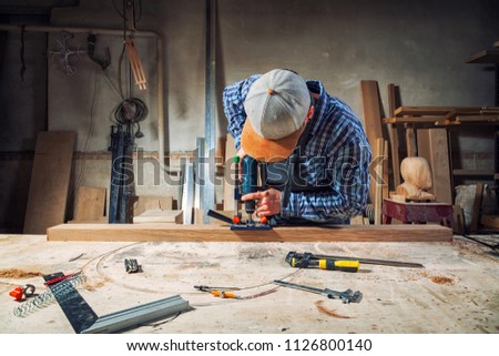 Experienced carpenter in work clothes and small buiness owner  is carving a wooden board on an  modern  hand drill in a light workshop side view, in the background a lot of tools