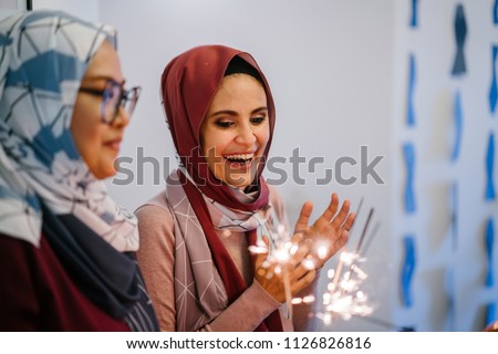 Two Muslim Malay woman celebrate their birthdays over a cake with sparklers on the cake. They are both wearing tudung hijab head scarves and smiling in joy.