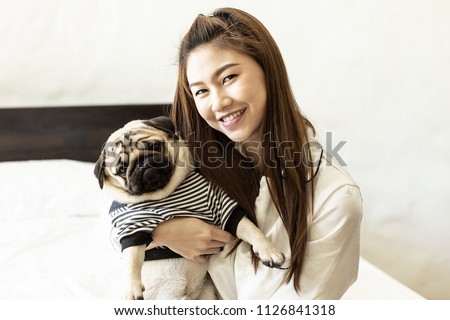Beautiful Attractive Asian young woman playing with her dog and smile with dog pug breed looking in funny face in bedroom,Friendship Concept