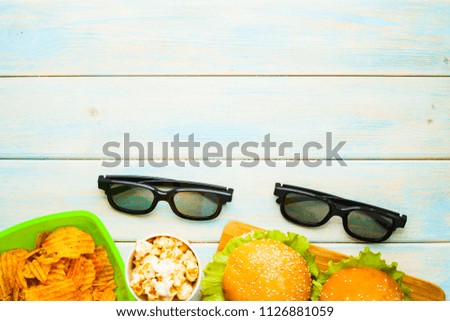 Weekend at home, leisure lifestyle, TV,  fast food concept. Set of 3D glasses, remote controllers and snacks in green bowls: chips, popcorn, burgers on the wooden background. Top view. Close up