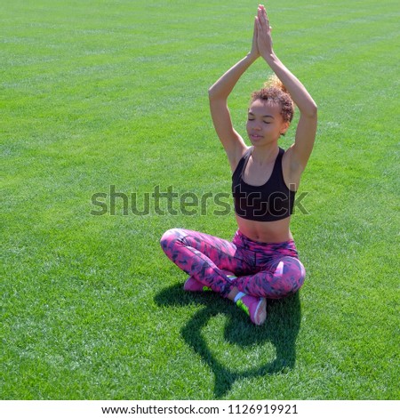 An African American girl in summer in sports clothes raised her hands up and meditates. Young cute black girl sitting on grass in relaxing pose with closed eyes. Square picture.