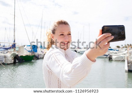 Portrait of beautiful young solo traveller tourist woman in coastal maritime port with boats destination, taking selfies photos with smartphone, smiling outdoors. Travel leisure recreation lifestyle.