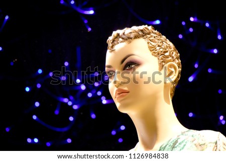 Close up of one female mannequin doll with short curly hair displayed n the shop window with blurred blue lights in the background , profile view