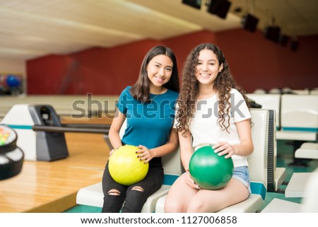 Female friends with bowling balls smiling while sitting on chairs in club