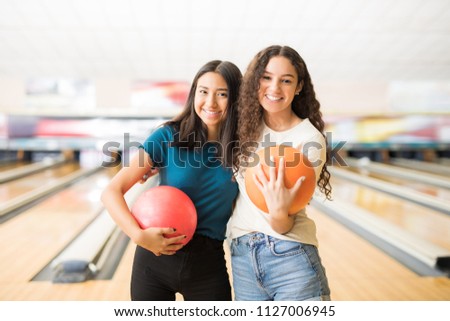 Smiling female friends with bowling balls hanging out in alley at club