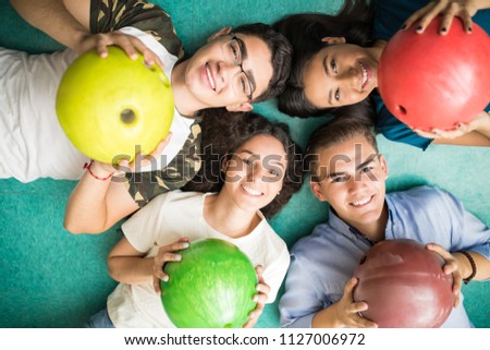 Directly above shot of multi-ethnic teenage friends showing colorful bowling balls while lying on alley at club
