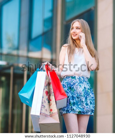 Lifestyle portrait young blonde girl, with shopping bags walking out from shop and talking by mobile phone.